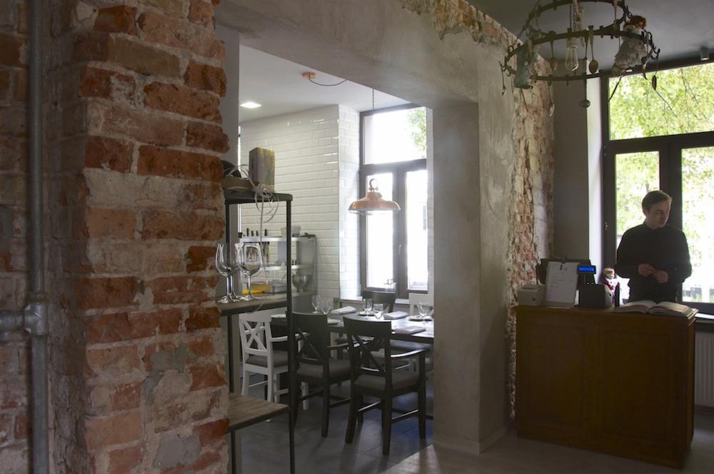 Wnętrze restauracji w stylu industrialnym z ceglaną ścianą, drewnianym stołem z nakryciem i kelnerem przy ladzie.