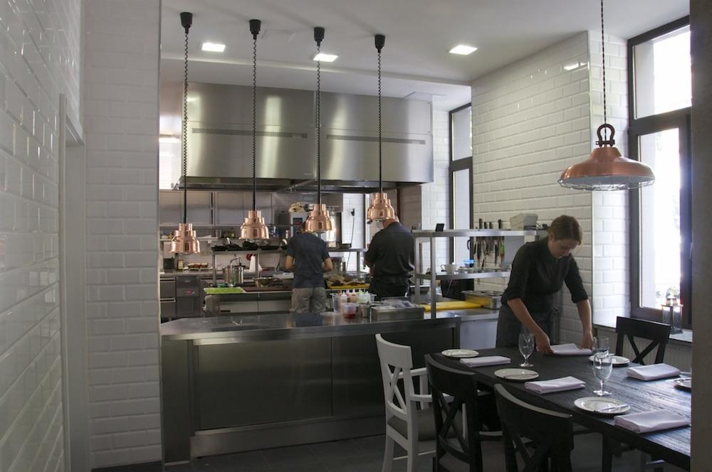 Restauracyjna kuchnia z widokiem na nakryty stół: dwie osoby pracują przy stalowych blatach pod miedzianymi lampami, trzecia osoba układa serwetki na ciemnym drewnianym stole z białymi krzesłami.