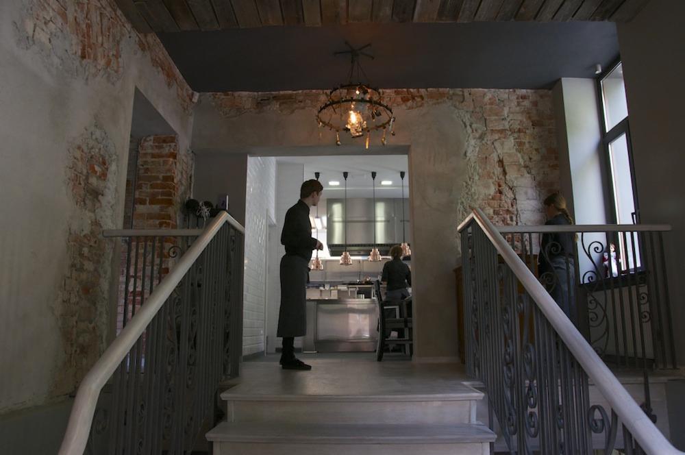 Wnętrze restauracji w Balicach, widok z góry schodów na otwartą kuchnię i obsługę, ściany z cegły i surowego tynku, ozdobny żyrandol.