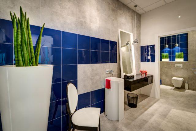 Nowoczesna łazienka z niebiesko-szarą mozaiką, wysoką białą donicą z zieloną rośliną, minimalistyczną umywalką z czerwonym ręcznikiem i czarną szafką z lustrem, uzupełniona figurką czerwonej krowy.