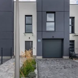 Nowoczesny dom bliźniak z szarą elewacją, oświetlony uliczną lampą LED, z podjazdem wyłożonym kostką brukową i minimalistycznym ogrodem z trawami ozdobnymi.