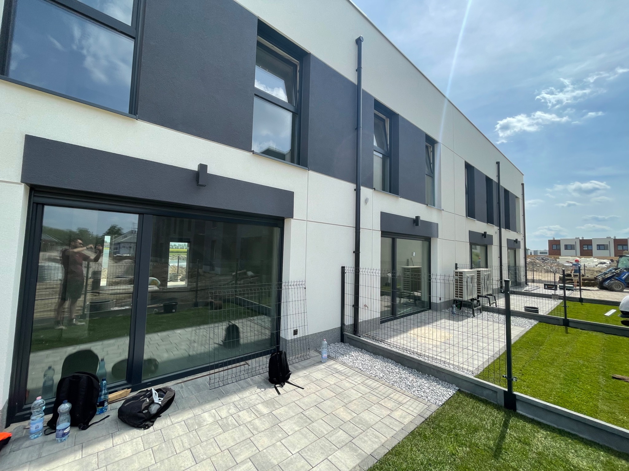 Nowoczesny dom w trakcie budowy, widoczne duże okna z czarnymi ramami, fragment elewacji w kolorze szarym i białym, teren wokół domu częściowo wyłożony kostką, częściowo trawa, w oknach odbija się...