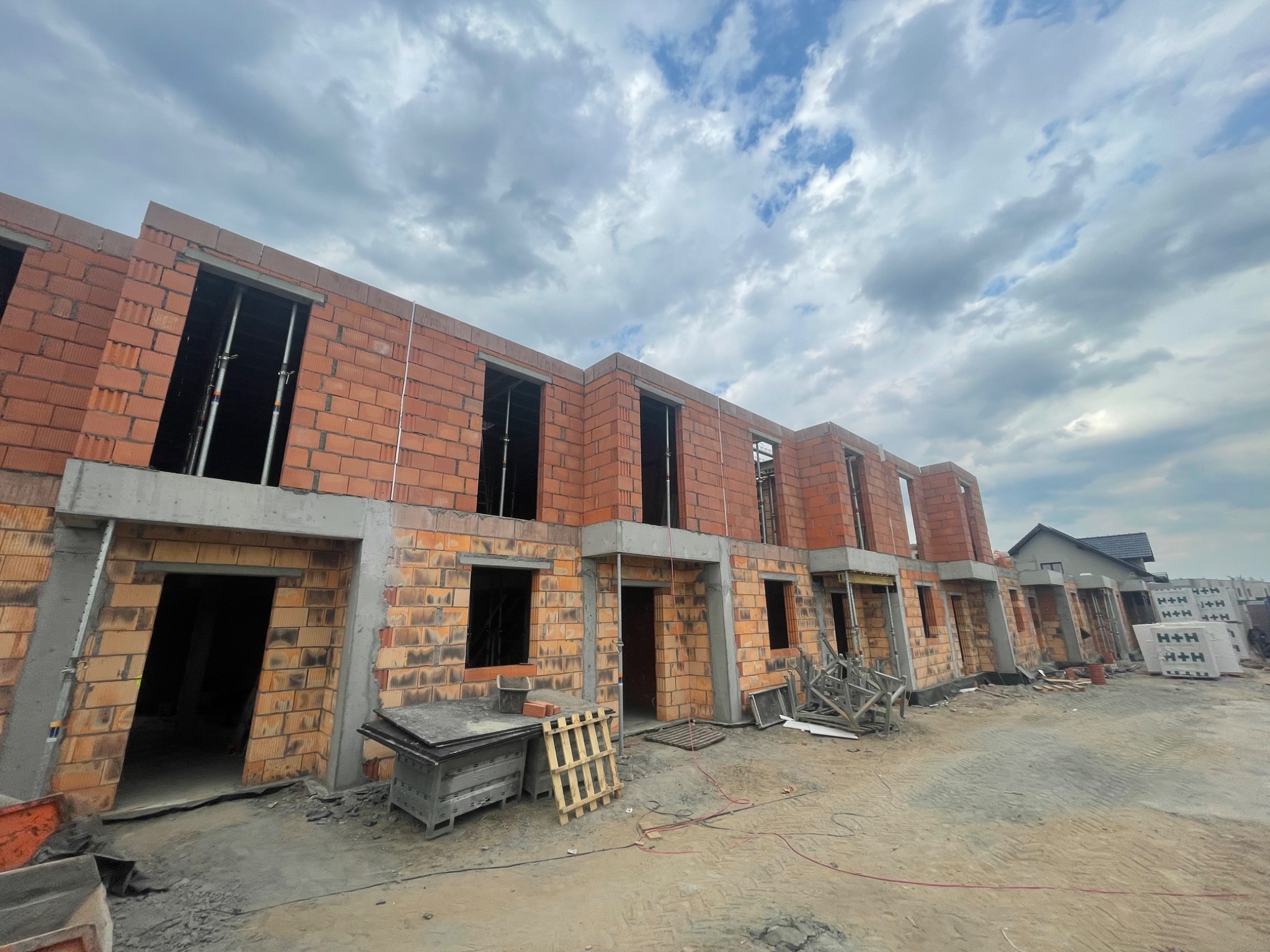Surowy stan szeregu domów w trakcie budowy z czerwonej cegły, widoczne pustaki H+H, niebo z chmurami w tle.