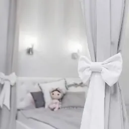 Jasnoszare zasłony z białymi kokardami w pokoju dziecięcym, łóżko z pluszową zabawką w tle.