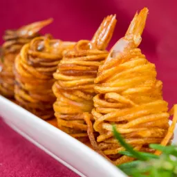 Elegancko podane krewetki owinięte smażonymi nitkami ziemniaczanymi na białym talerzu z dekoracją z rozmarynu na tle bordowej tkaniny.