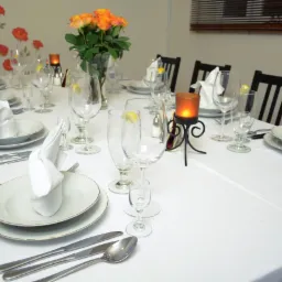 Elegancko nakryty stół z białym obrusem, talerzami, sztućcami, kieliszkami z plasterkiem cytryny, serwetkami ułożonymi w fantazyjne kształty, bukietem pomarańczowych róż i świecami w ozdobnych...