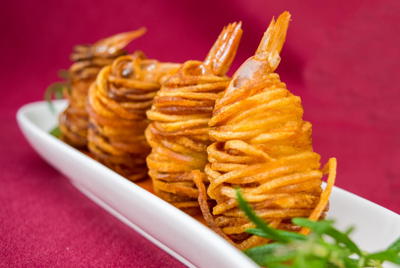 Elegancko podane krewetki owinięte smażonymi nitkami ziemniaczanymi na białym talerzu z dekoracją z rozmarynu na tle bordowej tkaniny.