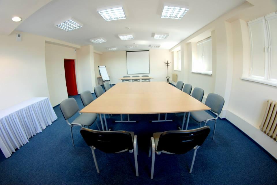 Sala konferencyjna z dużym stołem, krzesłami i projektorem, przygotowana do spotkania lub szkolenia.