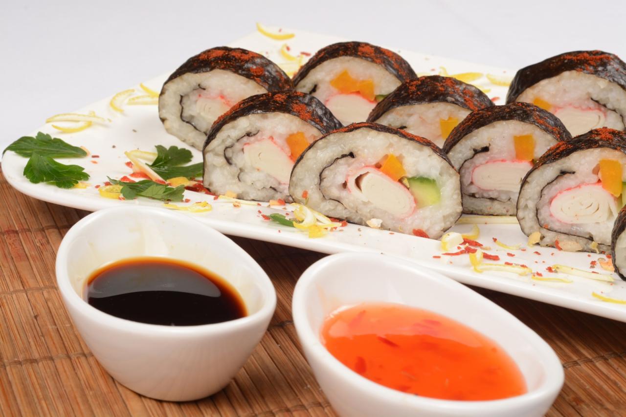 Talerz z sushi maki z krabem, ogórkiem i papryką, posypane przyprawą, podane z sosem sojowym i słodko-pikantnym na bambusowej podkładce.