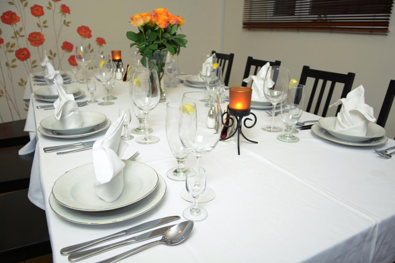 Elegancko nakryty stół z białym obrusem, talerzami, sztućcami, kieliszkami z plasterkiem cytryny, serwetkami ułożonymi w fantazyjne kształty, bukietem pomarańczowych róż i świecami w ozdobnych...