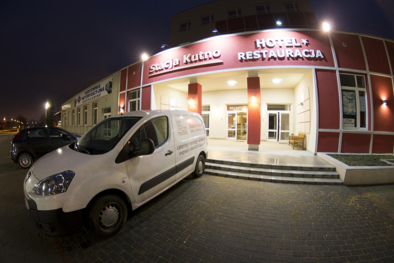 Biały samochód dostawczy z logo i napisem 'catering - noclegi - szkolenia - imprezy okolicznościowe' zaparkowany przed wejściem do hotelu i restauracji 'Stacja Kutno' nocą.