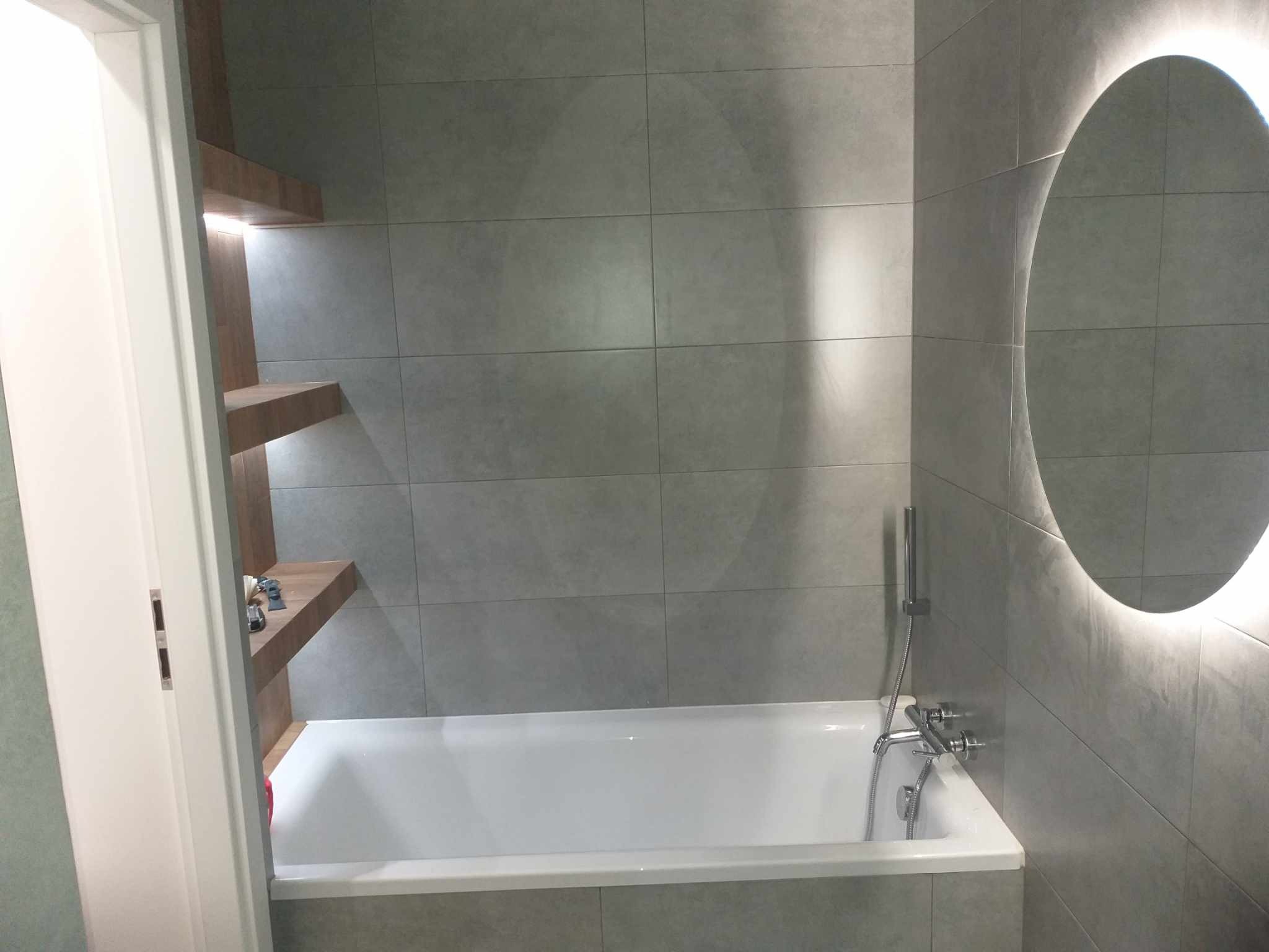 Nowoczesna łazienka z wanną w szarych płytkach, podświetlanym lustrem i drewnianymi półkami. Minimalistyczny design, oświetlenie LED, armatura chromowana.