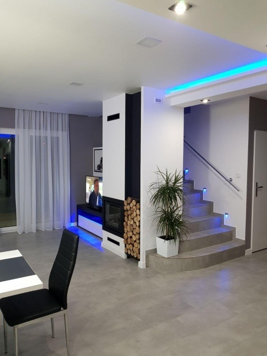 Nowoczesne wnętrze domu z kominkiem, drewnem, schodami z podświetleniem LED i minimalistycznym wystrojem. Widok na salon z krzesłem i telewizorem.