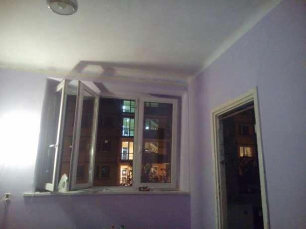 Pokój z fioletowymi ścianami, otwartym oknem z widokiem na oświetlony budynek nocą i białymi ramami okiennymi oraz białymi drzwiami po prawej stronie.