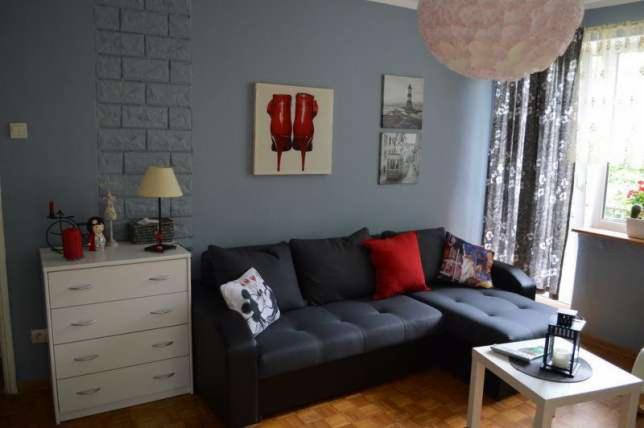 Szary narożnik z czerwoną poduszką w salonie z dekoracyjną ścianą imitującą cegłę i obrazem czerwonych szpilek.