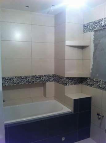 Wykończona łazienka z wanną obudowaną ciemnymi płytkami, jasne płytki na ścianach z dekoracyjnym pasem wzorzystych płytek, wnęki w ścianie wyłożone płytkami.