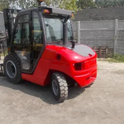 Czerwony wózek widłowy elektryczny Manitu z kabiną, stojący na szarym betonowym podłożu, z widocznym ogrodzeniem z betonowych płyt w tle.