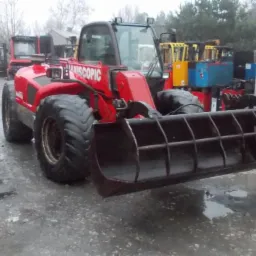 Czerwona maszyna budowlana z łyżką kratową z przodu, stojąca na mokrym placu, w tle inne maszyny i budynki.