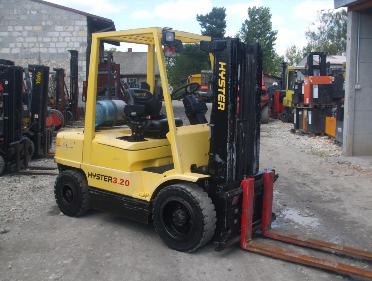Żółty wózek widłowy Hyster 3.20 z czarnym masztem i czerwonymi widłami, stojący na betonowym placu, z innymi wózkami widłowymi w tle.
