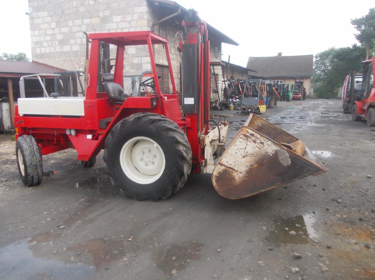 Czerwona maszyna z łyżką na nierównym, utwardzonym placu, w tle budynki i inne maszyny.