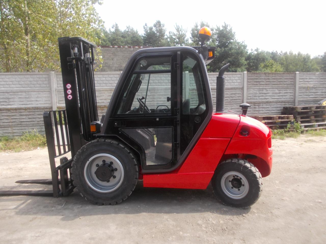 Czerwony wózek widłowy z kabiną, widok z boku na zewnątrz, z paletami w tle przy betonowym ogrodzeniu.