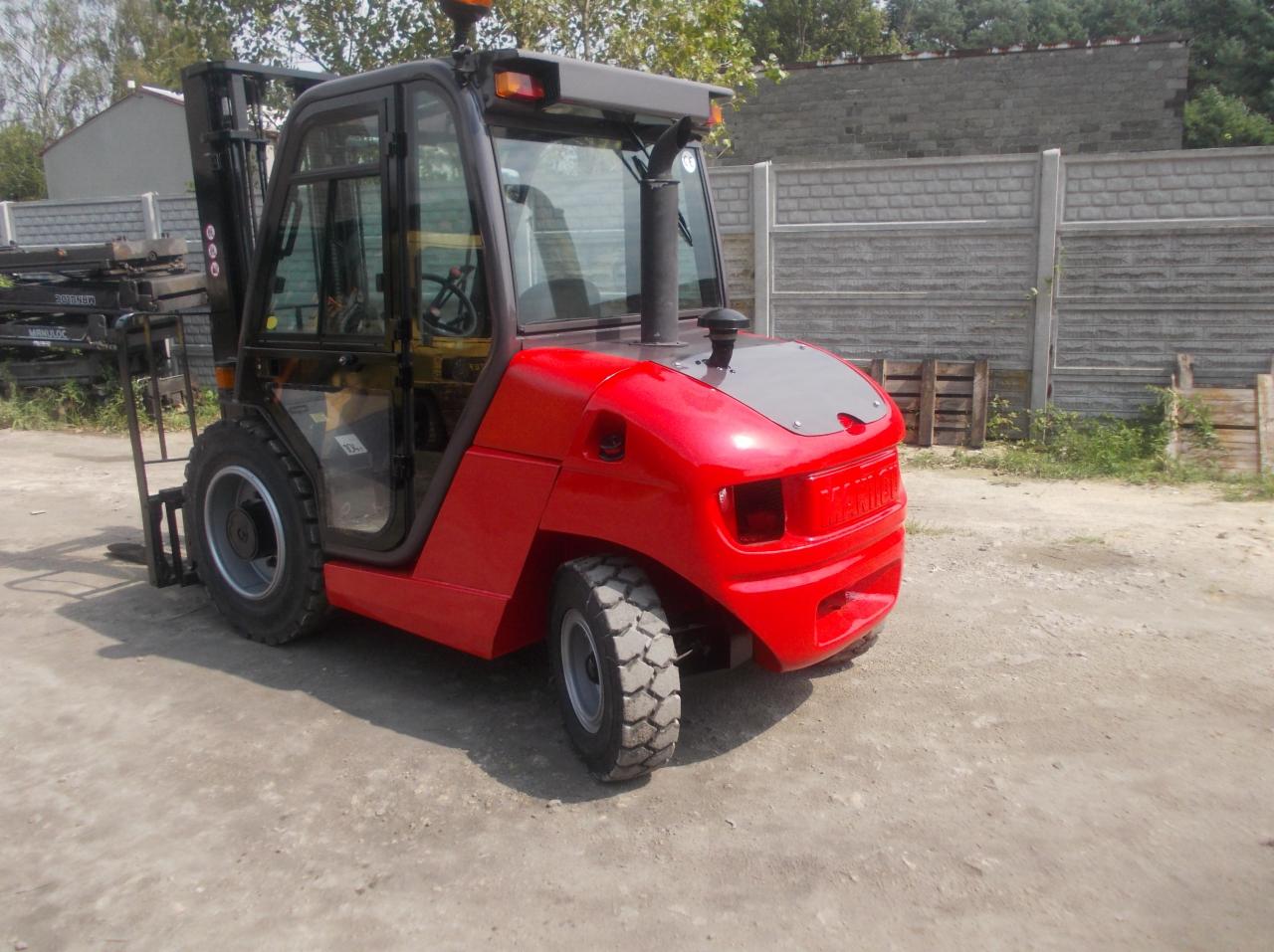 Czerwony wózek widłowy elektryczny Manitu z kabiną, stojący na szarym betonowym podłożu, z widocznym ogrodzeniem z betonowych płyt w tle.