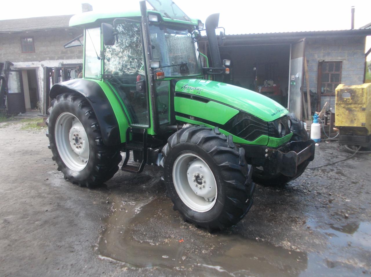 Zielony ciągnik Deutz-Fahr Agroplus 75 z kabiną, stojący na mokrym placu przed budynkiem gospodarczym z widocznym sprzętem warsztatowym w tle.