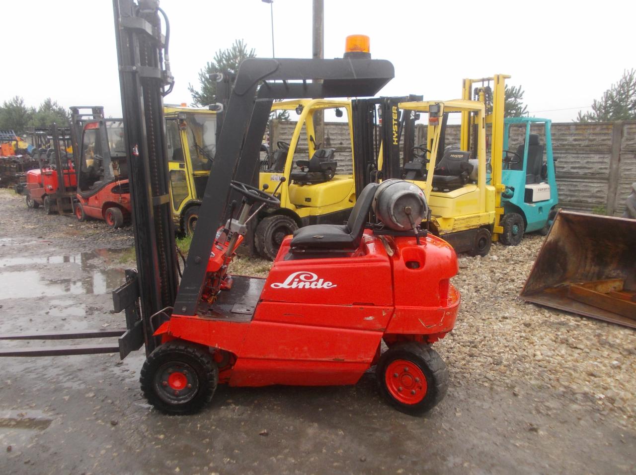 Czerwony wózek widłowy marki Linde na placu składowym, obok żółtych i niebieskich wózków widłowych innych marek, z widocznym zbiornikiem gazu na tylnej części.