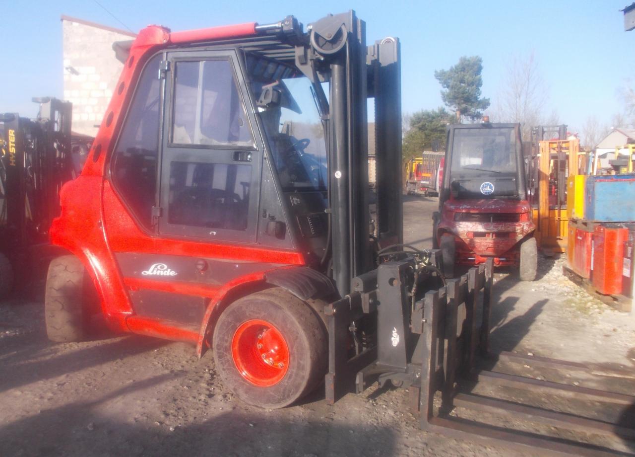 Czerwony wózek widłowy Linde z kabiną i czarnym masztem na szarym podłożu, drugi wózek widłowy w tle.