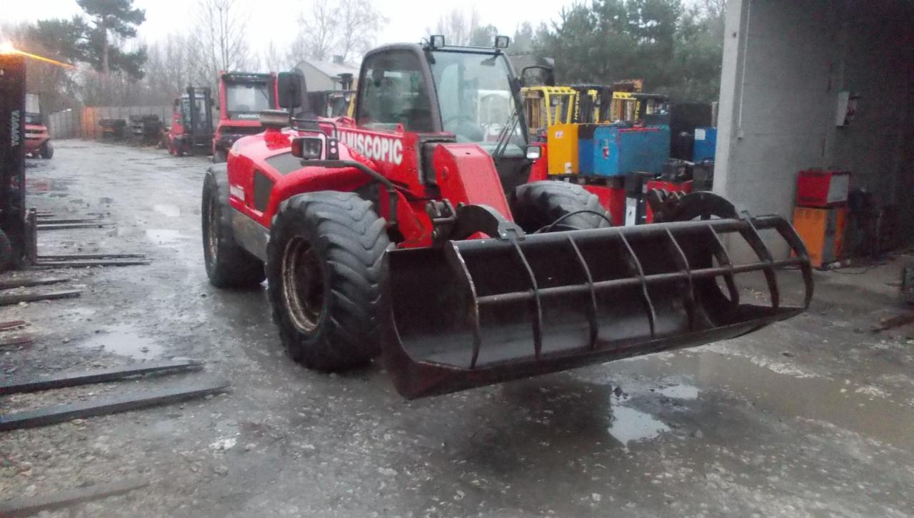 Czerwona maszyna budowlana z łyżką kratową z przodu, stojąca na mokrym placu, w tle inne maszyny i budynki.