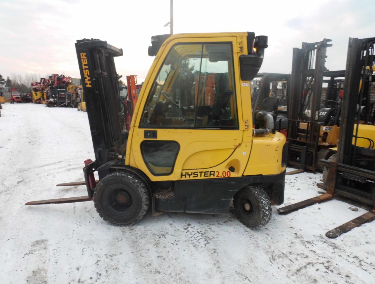 Żółty wózek widłowy Hyster 2.00 na śniegu, z innymi wózkami widłowymi w tle, widoczne ślady opon na śniegu.