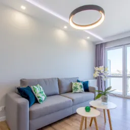 Jasnoszara sofa z dekoracyjnymi poduszkami w salonie z drewnianą podłogą, okrągłym wiszącym oświetleniem LED i białymi stolikami kawowymi.