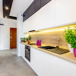 Nowoczesna kuchnia z białymi szafkami, drewnianym blatem, czarnym zlewem i roślinami doniczkowymi w kolorowych doniczkach, oświetlona listwą LED pod szafkami wiszącymi.