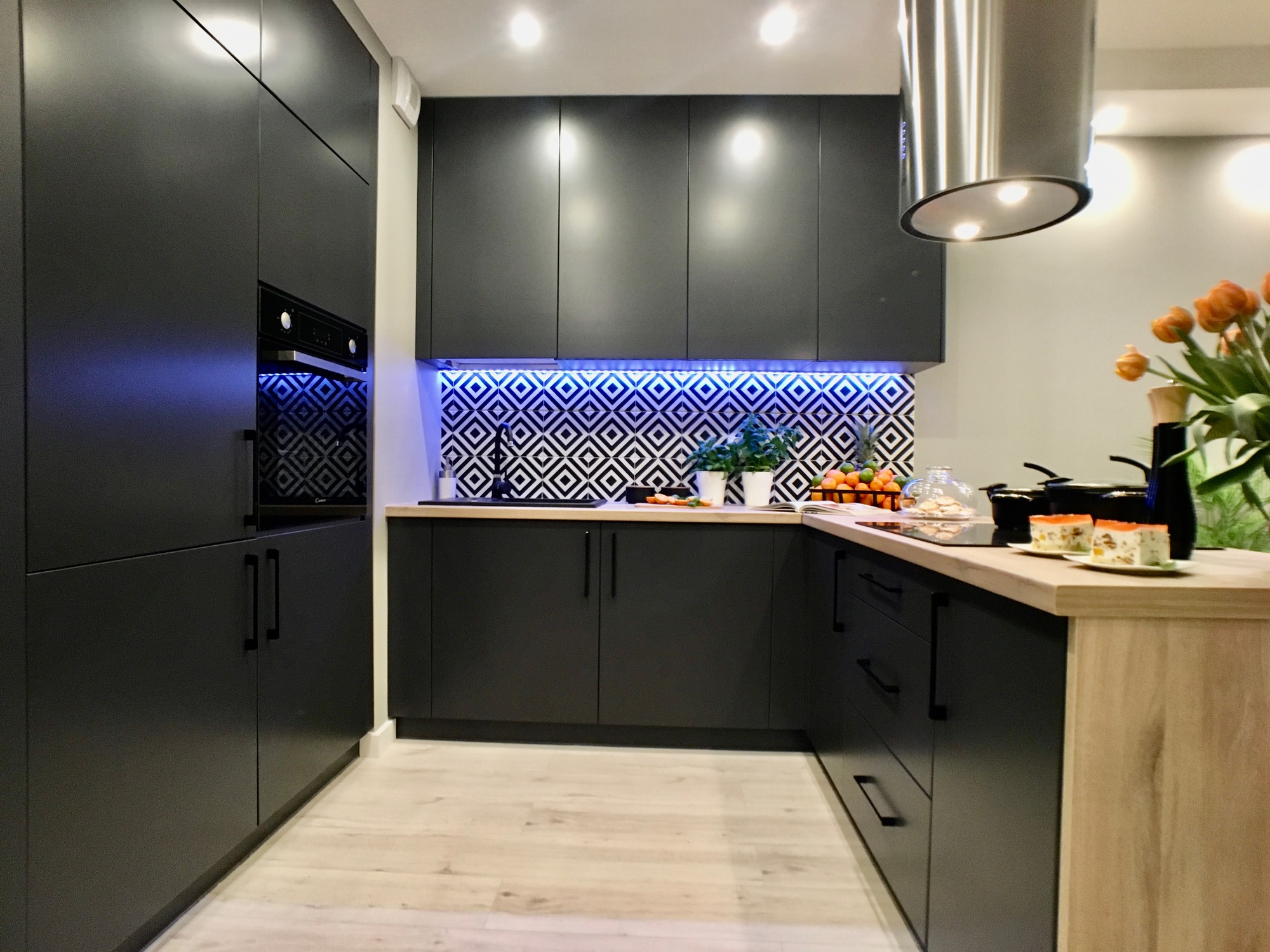 Nowoczesna kuchnia z czarnymi matowymi szafkami, drewnianym blatem, geometrycznymi płytkami podświetlonymi niebieskim światłem LED i stalowym okapem.