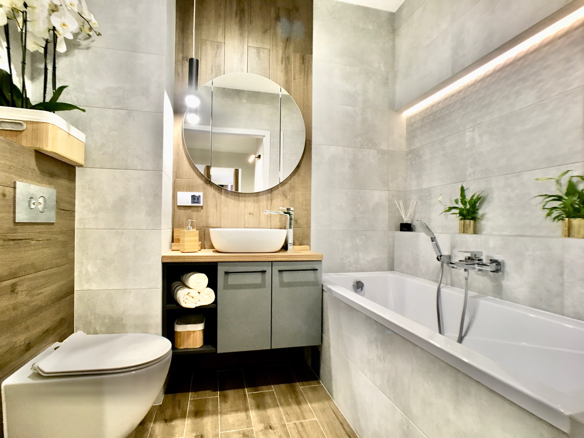 Nowoczesna łazienka z okrągłym lustrem, wiszącą umywalką, białą wanną i drewnopodobnymi płytkami na ścianie za toaletą.