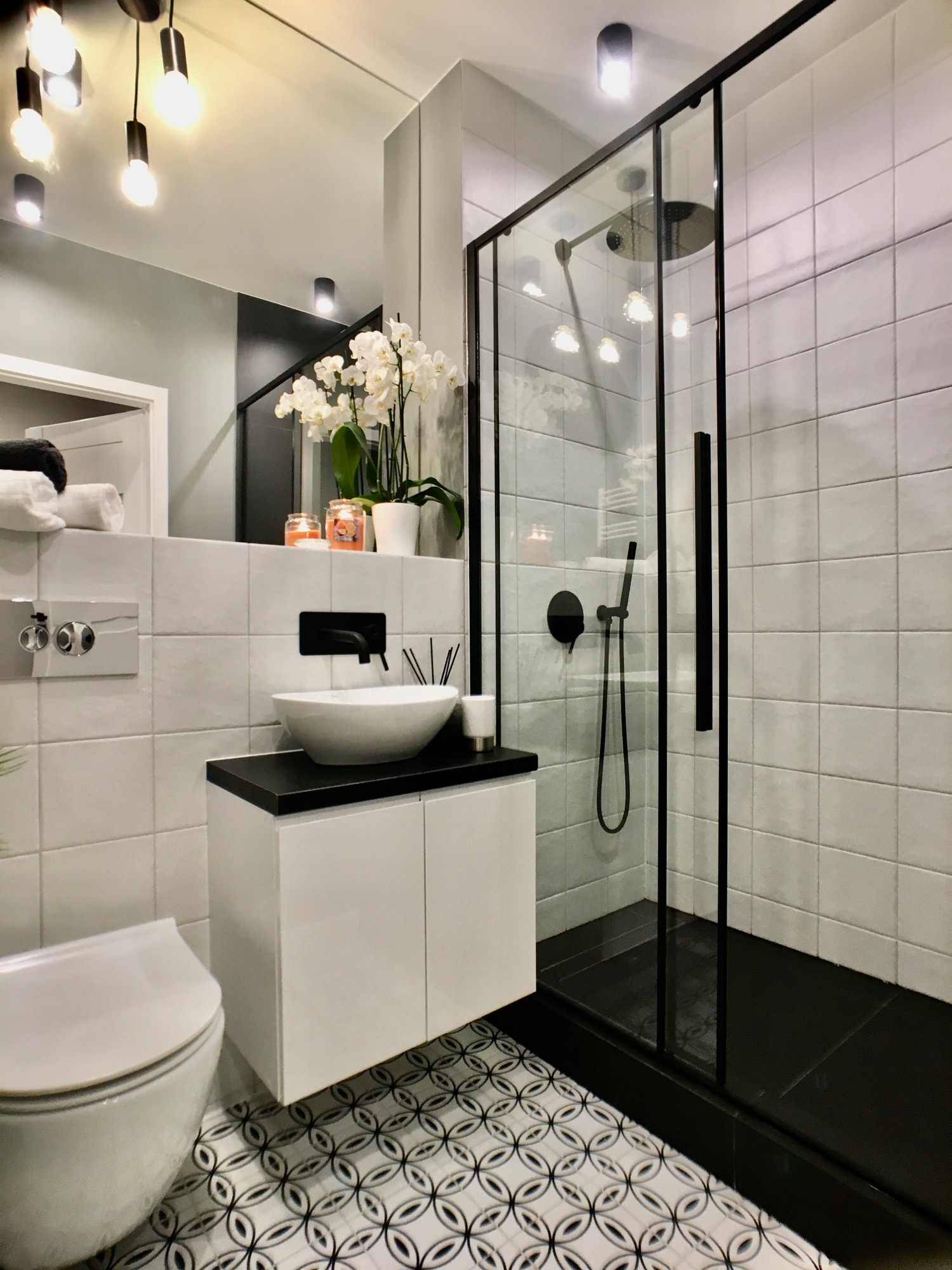 Nowoczesna łazienka w Siedlcach z białą armaturą, kabiną prysznicową z czarnymi profilami, wzorzystą podłogą i białymi płytkami na ścianach.