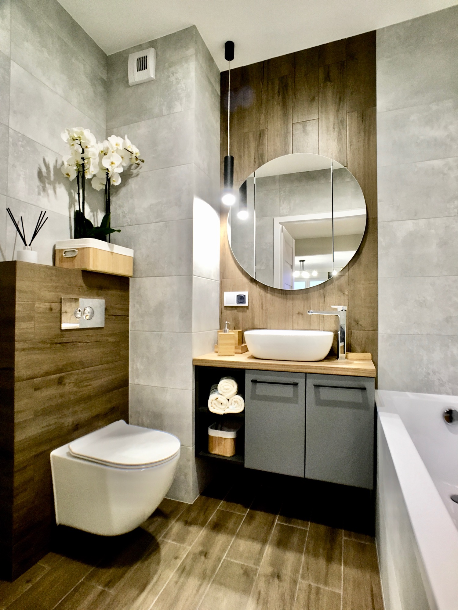 Nowoczesna łazienka z wiszącą miską WC, umywalką nablatową na drewnianym blacie i okrągłym lustrem podświetlonym lampą wiszącą, wykończona płytkami imitującymi beton i drewno.