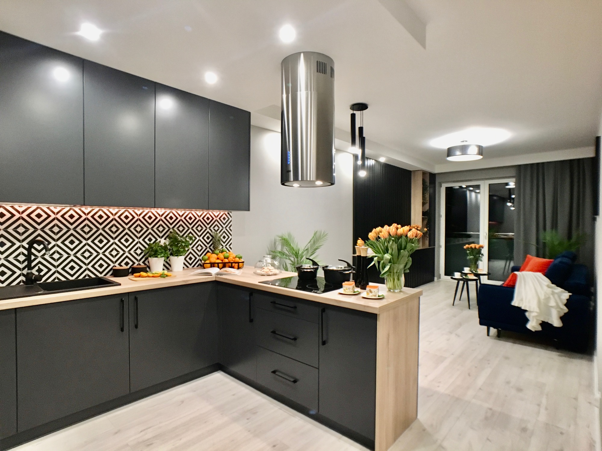 Nowoczesna kuchnia w odcieniach szarości z drewnianym blatem, geometryczne płytki nad blatem, stalowy okap cylindryczny, widok na salon z niebieską sofą i pomarańczowymi poduszkami.