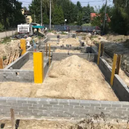 Wykop pod fundamenty z częściowo wzniesionymi ścianami z szarych bloczków betonowych, żółte szalunki, piasek wewnątrz, w tle drzewa i zabudowania.