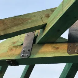 Konstrukcja drewnianego dachu z elementami stalowymi skręcana na śruby, widok od dołu na tle jasnego nieba.