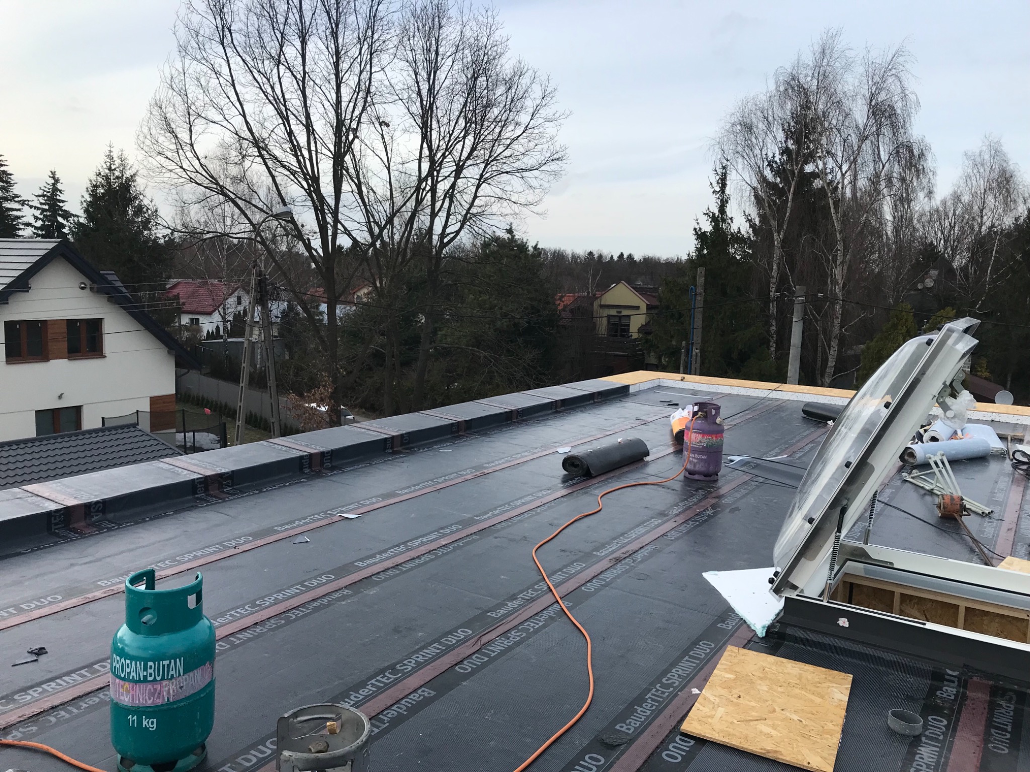 Montaż pokrycia dachowego z papy termozgrzewalnej na płaskim dachu, widoczne butle z gazem propan-butan, okno dachowe i narzędzia, w tle zabudowa jednorodzinna.