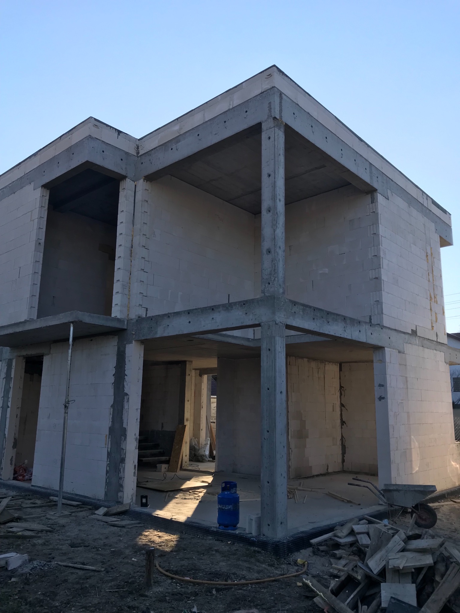 Surowy szkielet dwukondygnacyjnego domu z betonu komórkowego i betonowych słupów, widoczny niebieski cylinder gazowy i taczka na placu budowy.