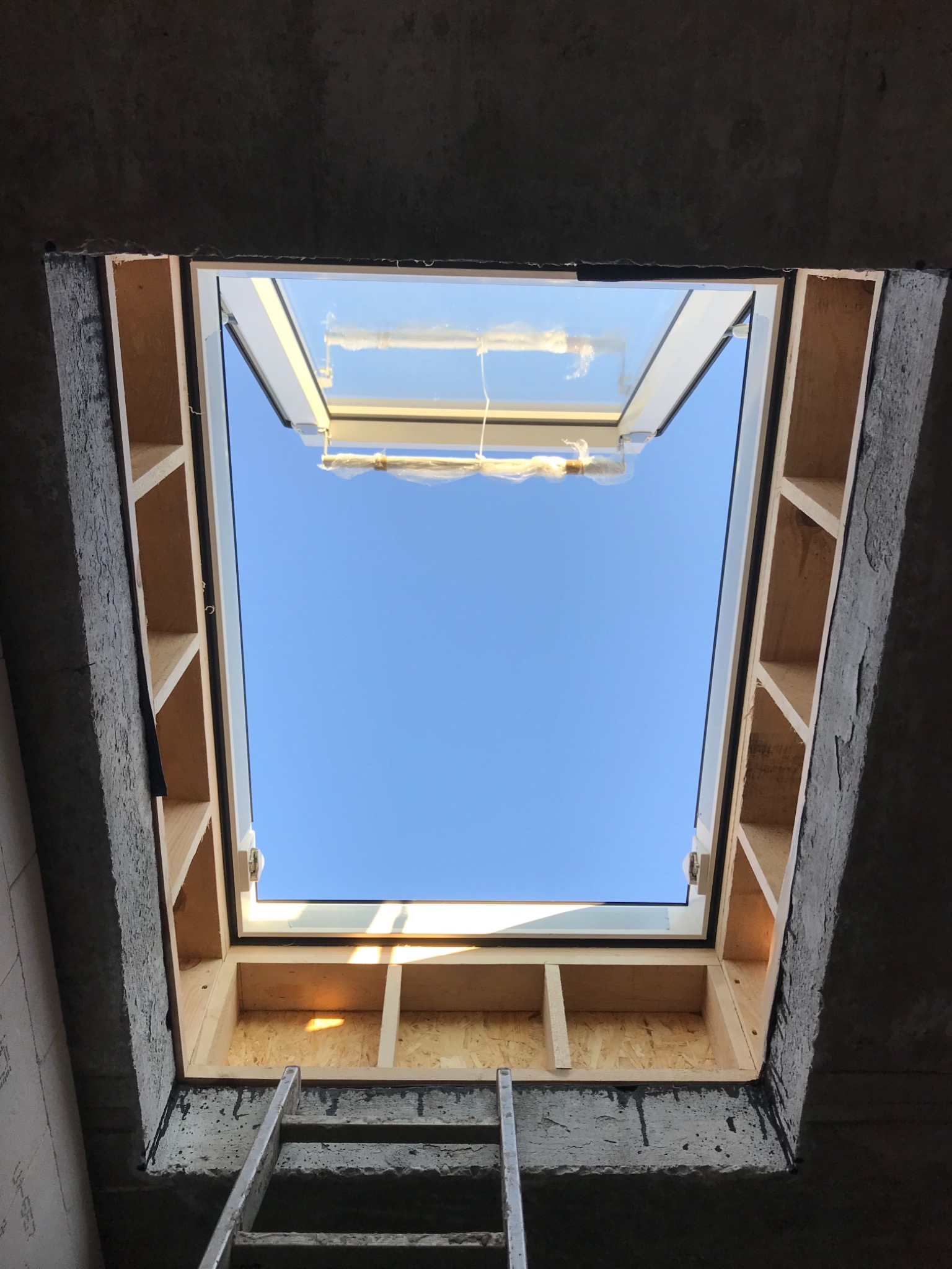 Montaż okna dachowego w surowym stanie, widoczne drewniane elementy konstrukcyjne i drabina oparta o betonowy otwór.