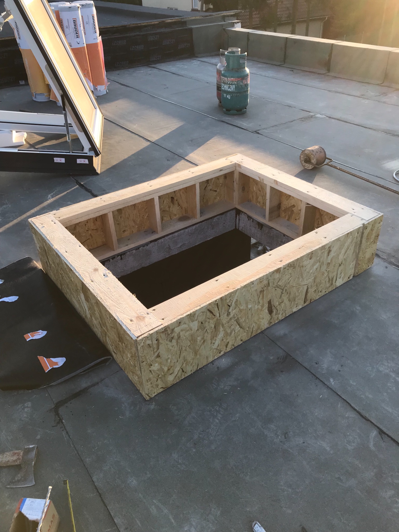 Otwór w dachu z drewnianą obudową z płyty OSB, przygotowany pod montaż okna dachowego; widoczne materiały izolacyjne w rolkach, butla gazowa i narzędzia.