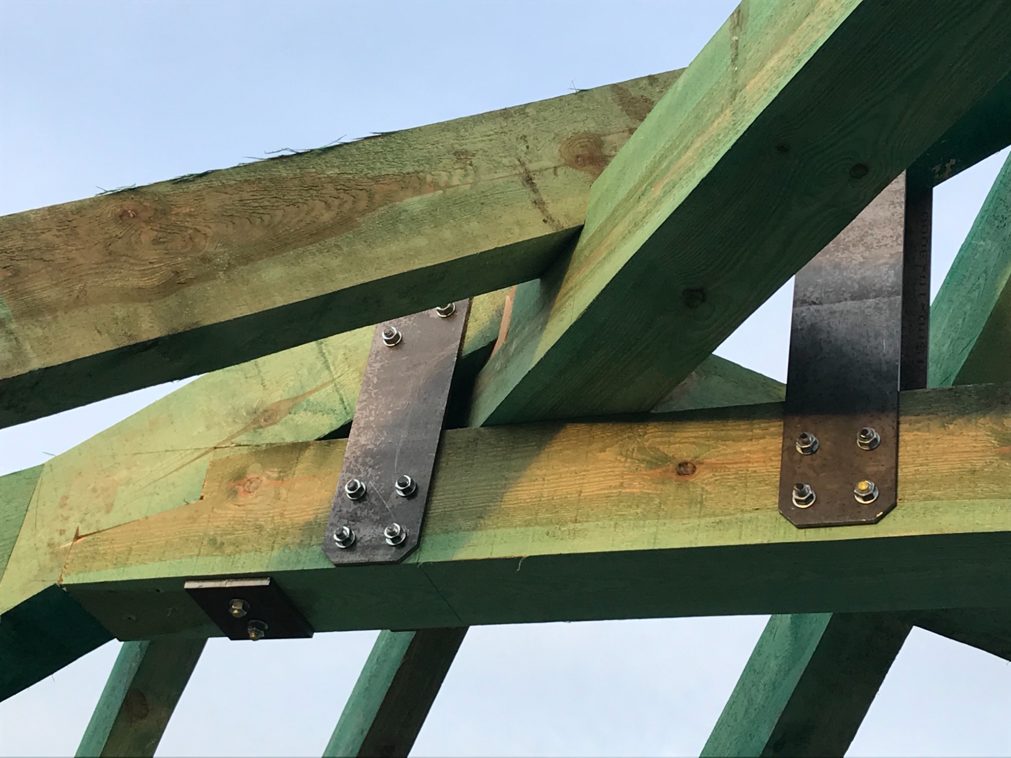 Konstrukcja drewnianego dachu z elementami stalowymi skręcana na śruby, widok od dołu na tle jasnego nieba.