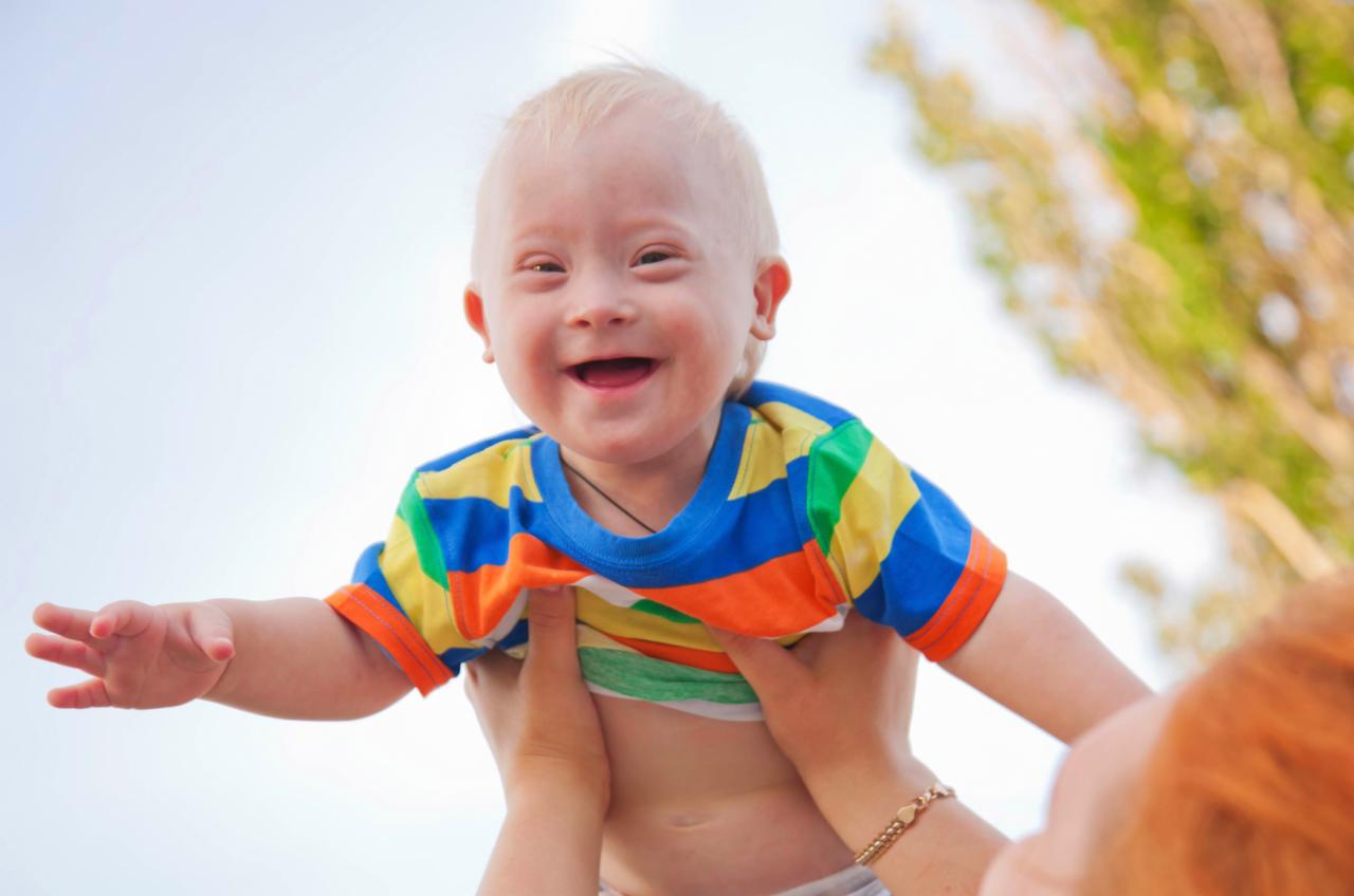 Uśmiechnięte dziecko z zespołem Downa, trzymane w górze przez osobę z rudymi włosami, na tle nieba i drzew.