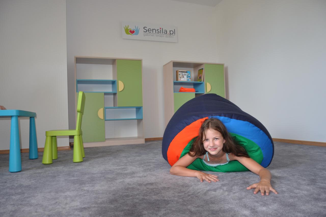 Dziewczynka uśmiecha się, leżąc w kolorowym tunelu sensorycznym w gabinecie terapii dzieci w Tarnowskich Górach, z widocznymi półkami na zabawki w tle.