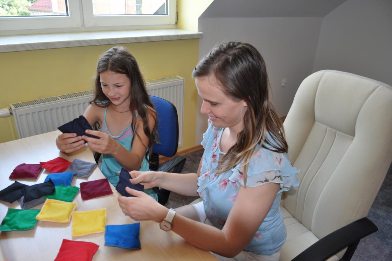 Dwie osoby siedzą przy stole, trzymając w rękach małe, kolorowe woreczki wypełnione materiałem. Na stole leżą inne woreczki w kolorach: czerwonym, niebieskim, żółtym, zielonym, szarym i bordowym.