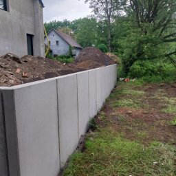 Marek Sosna - Nowo postawiony, szary mur z prefabrykowanych paneli betonowych, oddzielający teren z hałdami ziemi i gruzu od nierównego, trawiastego gruntu. W tle nowoczesny dom w budowie i stary budynek.