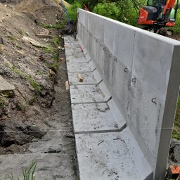 Marek Sosna - Montaż długiej, prefabrykowanej betonowej konstrukcji z metalowymi uchwytami w wykopie. Obok usypana ziemia, w tle mała koparka i drzewa.